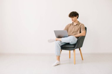 Beyaz duvara karşı otururken dizüstü bilgisayar kullanan çekici genç adam, kopyalama alanı. Milenyum erkeği çevrimiçi iletişim kuruyor, çalışıyor veya uzaktan taşınabilir bilgisayar üzerinde çalışıyor.