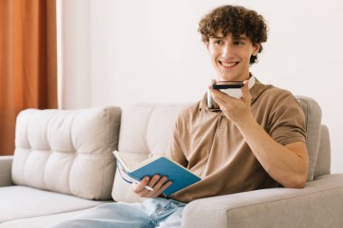 Genç, kıvırcık saçlı erkek öğrenci evde koltukta oturmuş sesli mesaj kaydediyor.