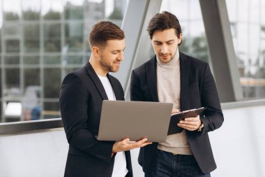 Takım elbiseli iki yakışıklı genç iş adamı bir dizüstü bilgisayar ve bir klasör tutuyorlar ve bir çalışma projesini tartışıyorlar ya da kentsel ofis binalarının arka planına karşı video görüşmesi yapıyorlar..