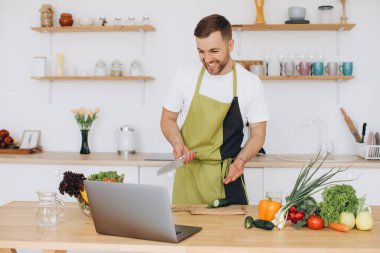 Evde mutlu bir adamın portresi, sebze salatası yapan bir adam kameraya bakıyor ve gülümsüyor, sebzeleri dilimliyor, dizüstü bilgisayarla çevrimiçi pişirme eğitimi alıyor.