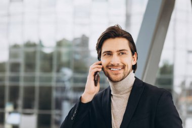 Takım elbiseli yakışıklı modern erkek işadamının portresi telefonla konuşuyor ve şehir binaları ve ofislerinin arka planında gülümsüyor..
