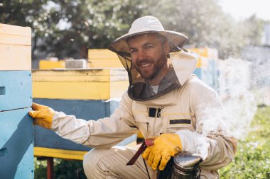 Arılarla arı kovanlarının yakınında çalışan mutlu bir erkek arı yetiştiricisinin portresi. Bal topla. Arı yetiştiricisi arıda. Arıcılık konsepti.