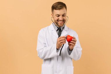 Erkek doktor kardiyolog bej arka planda steteskopla kalbi dinliyor.
