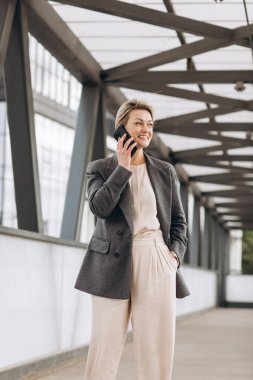 Takım elbiseli, gri ceketli, modern şehir ve ofis binalarında gülümseyen ve konuşan güzel bir iş kadınının portresi.
