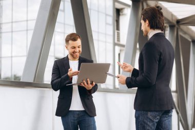 Laptop ve belge klasörlü iki modern mutlu iş adamı şehir ofislerinin ve binaların arka planına karşı bir şeyler tartışıyorlar.
