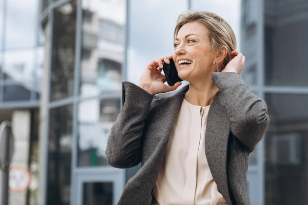 Portrait of a beautiful mature business woman in suit and gray jacket smiling and talking on a phone on the modern urban and office buildings background