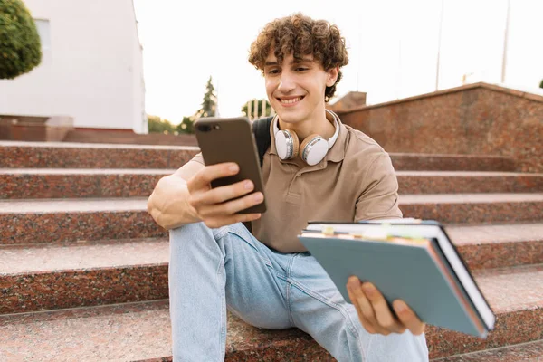 魅力的な巻き毛の若い男の大学や大学生が階段に座ってスマートフォンを見て — ストック写真
