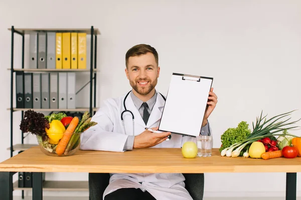 新鮮な野菜の間でオフィスのデスクで職場に座っている幸せな医師の栄養士とコピースペース 食事計画のコンセプトとフォルダを表示 — ストック写真