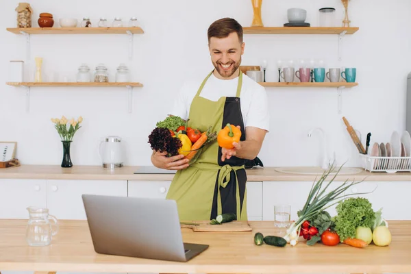 stock image Portrait of happy man at home, man cooking vegetable salad looking at camera and smiling, slicing vegetables, using laptop for online cooking training