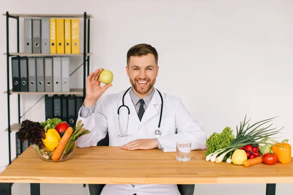新鮮な野菜の間で職場のテーブルに座っている幸せな医師栄養士とリンゴを保持 — ストック写真