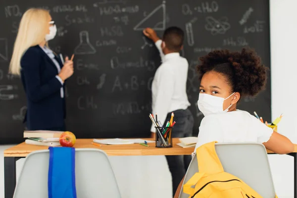 Portrait of African American school girl in mask sitting at desk on a blackboard background in a classroom during a lesson. Mask mode, quarantine, covid-19.