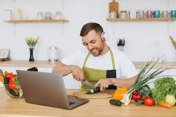 stock image Happy man in headphones preparing salad in kitchen from fresh vegetables and looking at laptop.