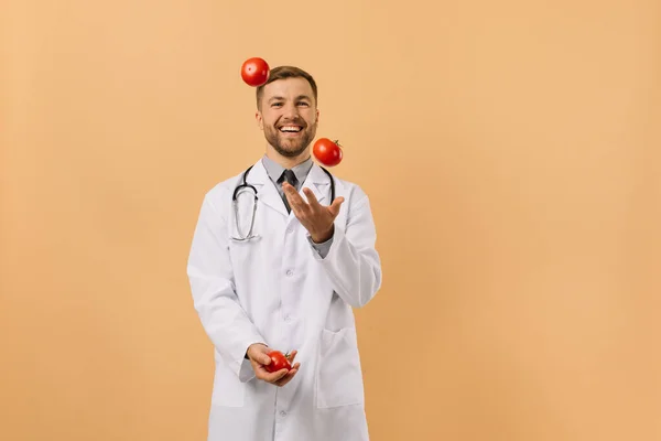Άνδρας Διατροφολόγος Γιατρός Στηθοσκόπιο Χαμογελώντας Και Ζογκλέροντας Ντομάτες Μπεζ Φόντο — Φωτογραφία Αρχείου