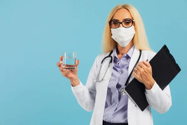 Mature woman doctor in mask holds a glass of water and recommends drinking water on blue background. Covid-19 concept