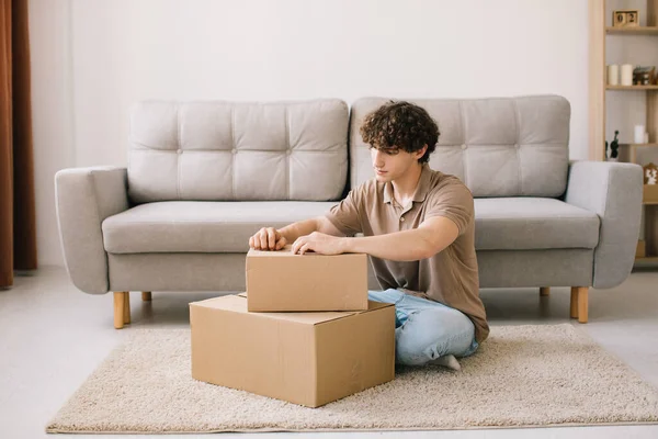 Happy young smiling curly man opening box with ordered goods gifts, presents at home on couch. Online shopper male customer opening online shop parcel.