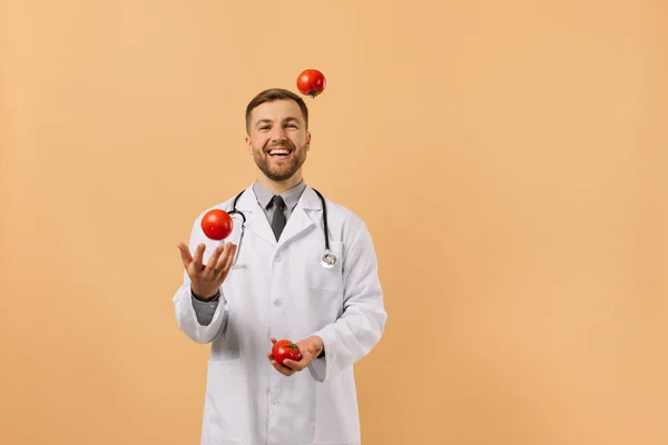 Médico Nutricionista Masculino Com Estetoscópio Sorrindo Malabarismo Tomates Fundo Bege — Fotografia de Stock