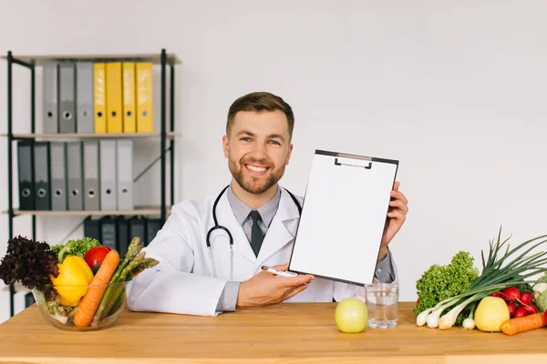 Happy doctor nutritionist sitting at workplace at desk in office among fresh vegetables and showing folder with copy space, diet plan concept