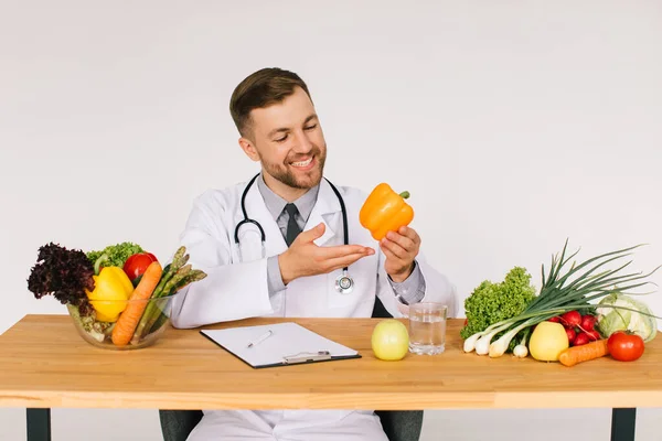 Happy doctor nutritionist sitting at workplace at desk in office among fresh vegetables and holding pepper, diet plan concept