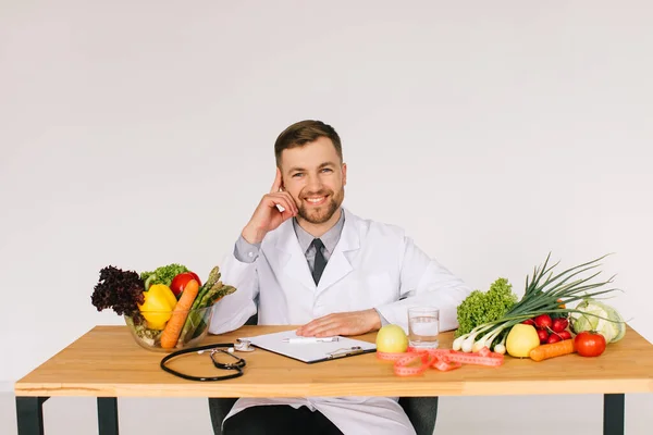 Happy doctor nutritionist sitting at workplace at desk in office among fresh vegetables, diet plan concept