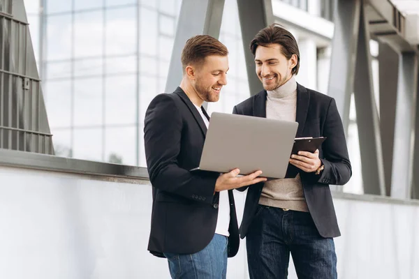 stock image Two handsome young male businessmen in suits hold a laptop and a folder with documents and discuss a working project or have a video call against the background of urban office buildings.