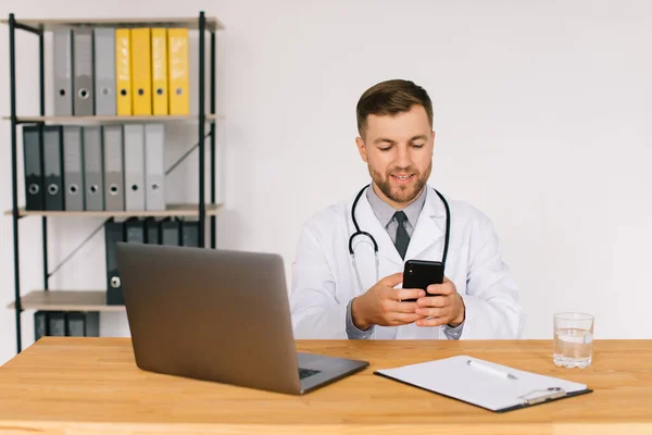 stock image Male doctor holds a phone and sits in the office near the laptop