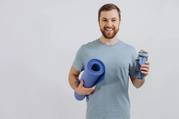 stock image Portrait of handsome bearded man in sportswear holding yoga mat and bottle of fresh water on white background, copy space