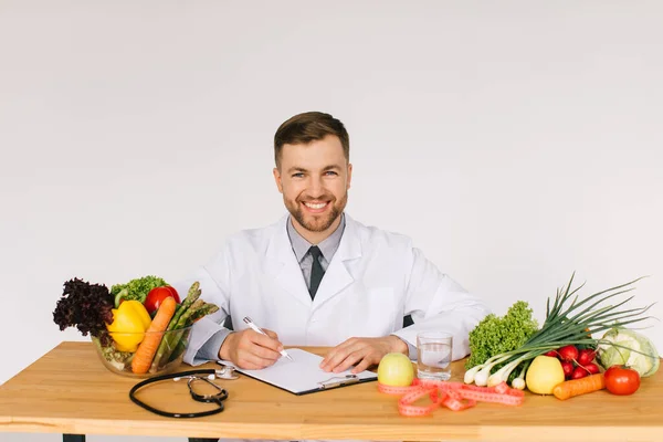 Happy doctor nutritionist sitting at workplace at desk in office among fresh vegetables, diet plan concept