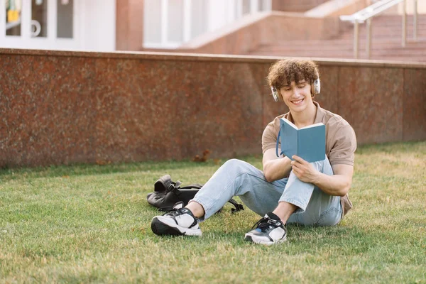 草の上に座って大学の近くに勉強ヘッドフォンで魅力的な男性学生 試験の準備 — ストック写真