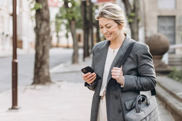 Gri Ceketli Olgun Sarışın Bir Kadını Gülümsüyor Elinde Telefonla Şehirde — Stok fotoğraf