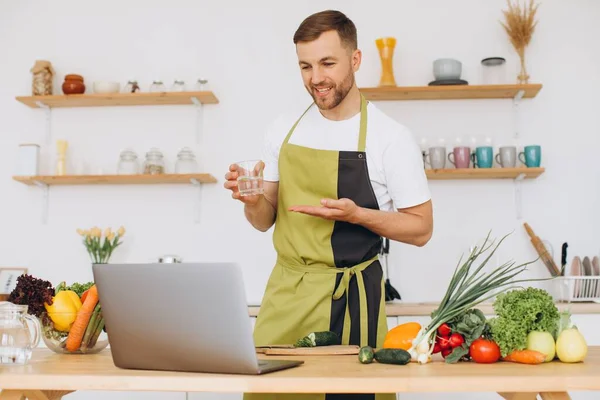 stock image Portrait of happy man at home, man cooking vegetables and showing glass of water, slicing vegetables, using laptop for online cooking training