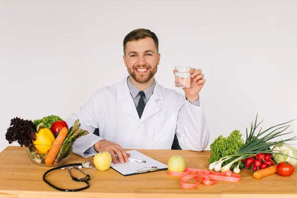 快乐的医生营养师坐在办公室的办公桌前 一边吃着新鲜蔬菜一边喝着一杯水 一边制定了饮食计划 — 图库照片