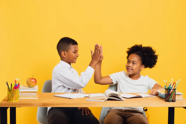 stock image Happy african american schoolgirl and schoolboy sitting together at desk and giving high five to each other on yellow background. Back to school concept.