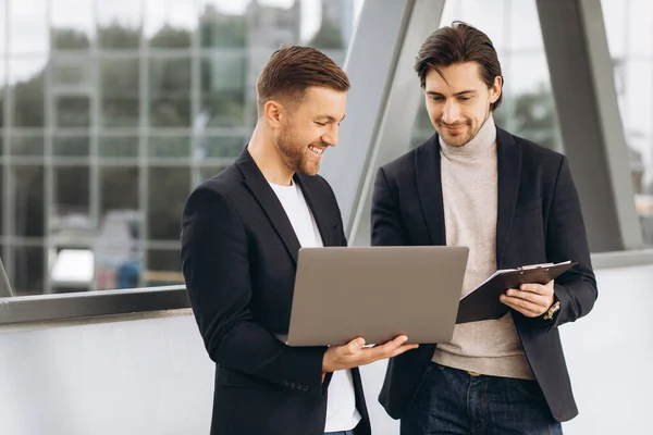 stock image Two handsome young male businessmen in suits hold a laptop and a folder with documents and discuss a working project or have a video call against the background of urban office buildings.