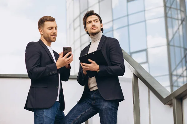 stock image Two modern happy businessmen with phone and folder of documents discussing something against the background of urban offices and buildings