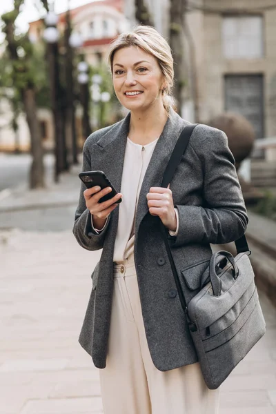 熟女ブロンドビジネス女性でAグレージャケット笑顔で街を歩き 電話を保持 — ストック写真