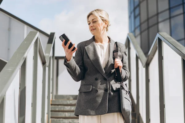一个穿着西服和灰色夹克的漂亮而成熟的女商人的画像 在现代都市背景下微笑着和电话交谈 — 图库照片