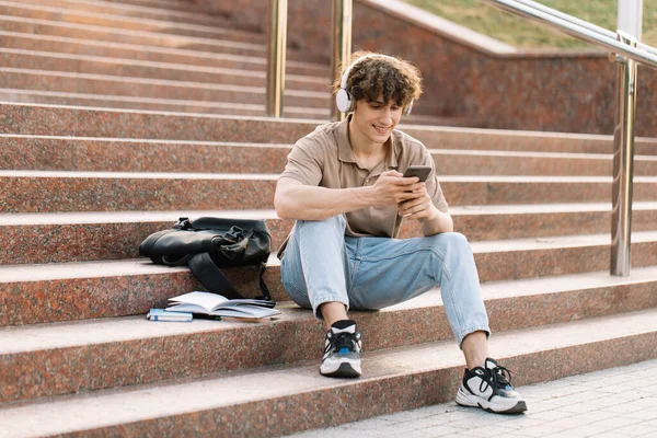 魅力的な巻き毛の若い男の大学や大学生が階段に座ってスマートフォンを見て — ストック写真