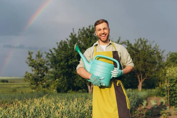 快乐的男性农民在花园里拿着一个浇水罐 背靠着雨后的夏色彩虹 — 图库照片