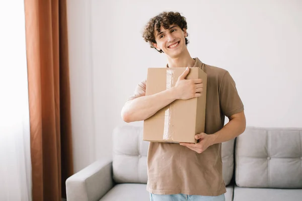 Shopping And Delivery. Curly Young Man Customer Holding Cardboard Box Posing Smiling To Camera Standing At Home. Excited Male Buyer Hugging Delivered Parcel. Shopaholism Concept