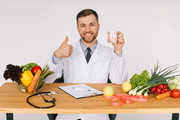 Happy doctor nutritionist sitting at workplace at desk in office among fresh vegetables holding glass of water, diet plan concept