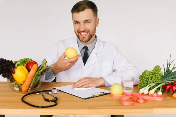 Happy doctor nutritionist sitting at workplace table among fresh vegetables and holding apple