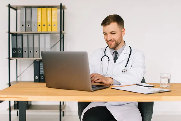 職場でノートパソコンを持って座っている間 幸せな男性医師は オンライン医療のウェビナーやトレーニングセミナーを見て — ストック写真