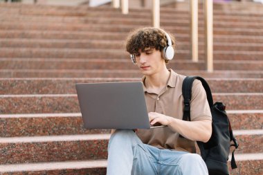 E-öğrenme, uzaktan öğrenme veya uzaktan öğrenme kavramı, üniversitede basamaklarda dizüstü bilgisayar kullanan genç kıvırcık saçlı okul çocuğu, üniversite ya da üniversite öğrencisi