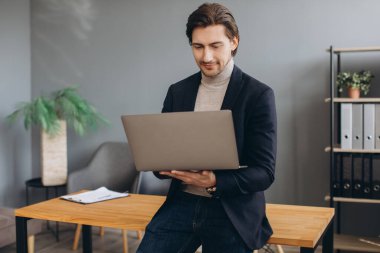 Modern ofiste takım elbiseli ve dizüstü bilgisayar kullanan genç ve neşeli iş adamının görüntüsü