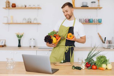 Evde mutlu bir adamın portresi, sebze salatası yapan bir adam kameraya bakıyor ve gülümsüyor, sebzeleri dilimliyor, dizüstü bilgisayarla çevrimiçi pişirme eğitimi alıyor.