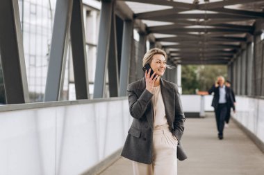 Takım elbiseli, gri ceketli, modern şehir ve ofis binalarında gülümseyen ve konuşan güzel bir iş kadınının portresi.