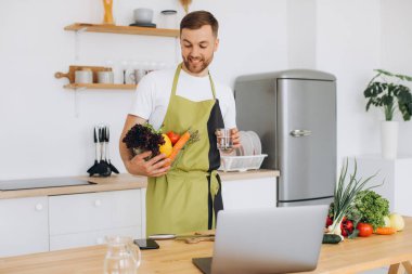 Evde mutlu bir adamın portresi, sebze salatası yapan bir adam kameraya bakıyor ve gülümsüyor, sebzeleri dilimliyor, dizüstü bilgisayarla çevrimiçi pişirme eğitimi alıyor.