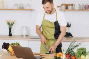 Evde mutlu bir adamın portresi, sebze salatası yapan bir adam kameraya bakıyor ve gülümsüyor, sebzeleri dilimliyor, dizüstü bilgisayarla çevrimiçi pişirme eğitimi alıyor.