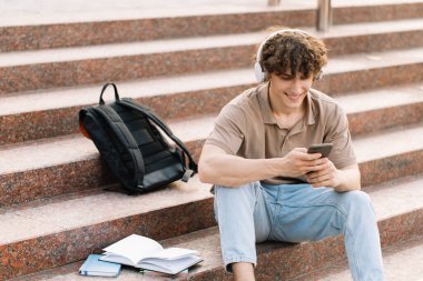 Çekici kıvırcık saçlı genç adam üniversite ya da üniversite öğrencisi merdivenlerde oturuyor ve akıllı telefon arıyor.