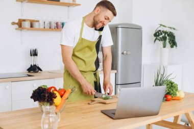 Çekici genç adam mutfakta dizüstü bilgisayarla yemek pişiriyor ve akıllı telefondan konuşuyor..